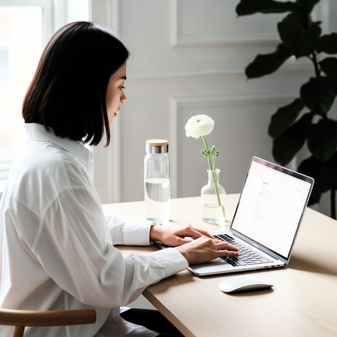 Woman working on a laptop at a tidy desk with a water bottle and a single white flower in a vase.