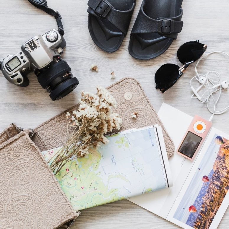 Travel flat lay featuring camera, map, sandals, sunglasses, flowers, and pouch.