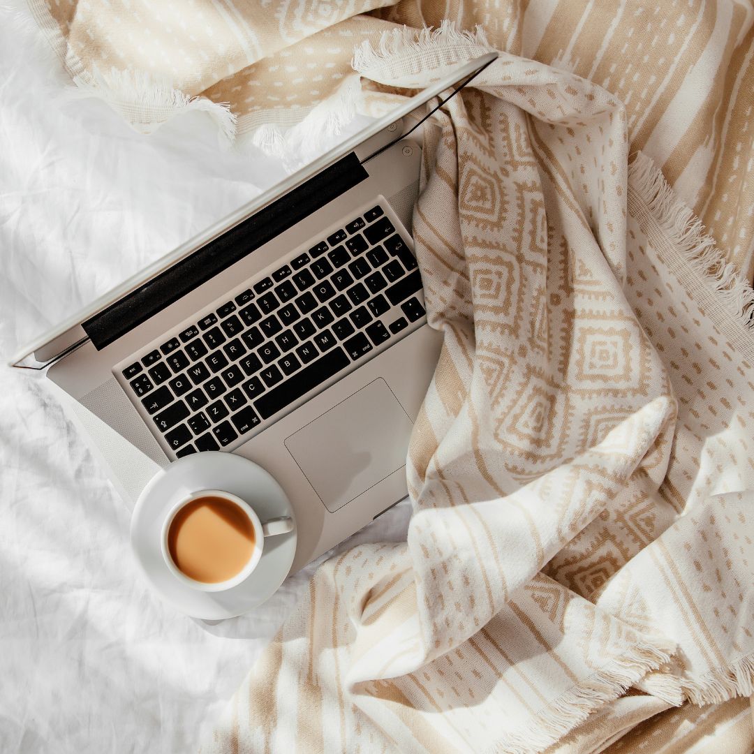 A laptop and a cup of coffee on a white bed with a beige patterned blanket